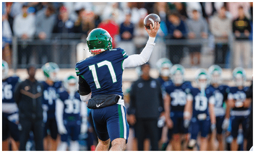 A person wearing a green helmet and blue sports uniform with the number 17 is preparing to throw a football on a field with a blurred crowd in the background. Description generated by AI