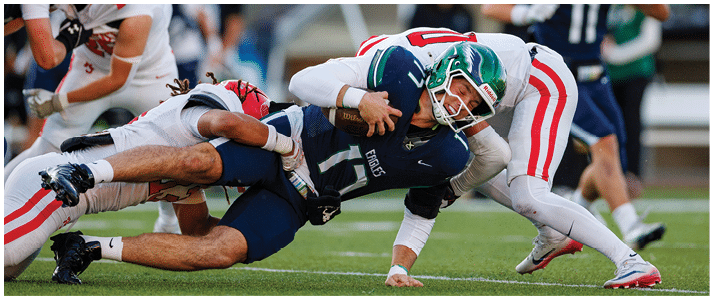 People in football uniforms are engaged in a tackle during a game on a field with blurred spectators in the background. Description generated by AI
