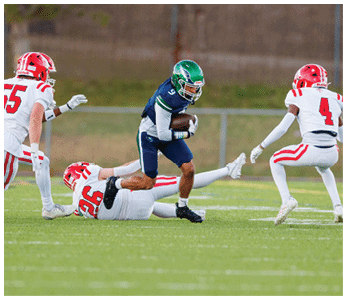 A person in a blue and green football uniform is running with the ball while three people in white and red football uniforms attempt to tackle or block them on a grassy field. Description generated by AI