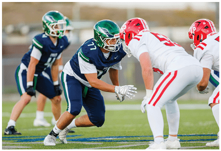 Two people in blue and green football uniforms are positioned opposite two people in white and red football uniforms on a grassy field during a game. Description generated by AI