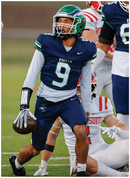 A person wearing a green helmet and navy blue football uniform with the number nine is holding a football and appears to be preparing to throw it while another person in a white and red uniform is on the ground behind them on a football field. Description generated by AI