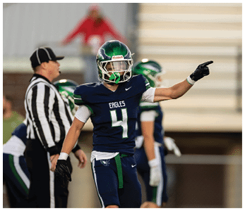 A person wearing a dark blue football uniform with the number 4 and the word Eagles is pointing while standing on a football field with a referee and other players in the background. Description generated by AI