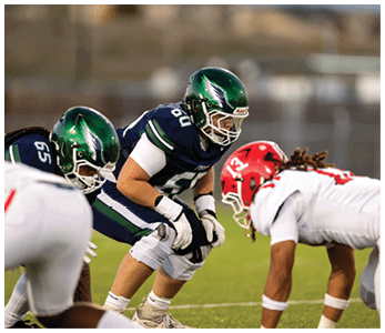 Two football players in dark green and blue uniforms face off against two players in white uniforms on a grassy field during a game. Description generated by AI