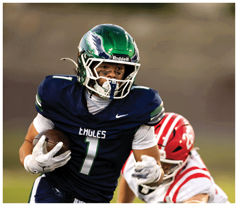 A person wearing a green helmet and navy blue football uniform with the word Eagles is running with a football while another person in a red and white uniform attempts to catch them. Description generated by AI