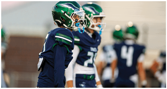 People wearing dark blue football uniforms and green helmets stand on a field, with one person in the foreground looking to the side. Description generated by AI