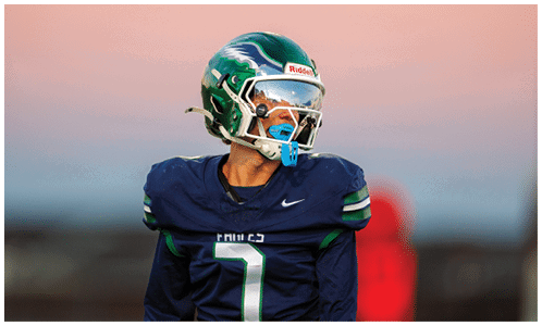 A person wearing a dark blue football uniform and green helmet is looking to the side against a blurred background with a gradient sky. Description generated by AI