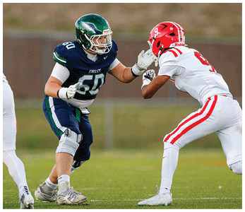 Two people in football uniforms are facing each other on a field, one wearing a dark blue and green uniform and the other in a white and red uniform, both in a ready stance. Description generated by AI