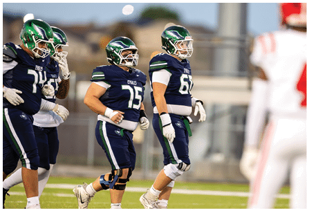 Four people in dark blue football uniforms and green helmets are walking on a field while a person in a white and red uniform stands nearby. Description generated by AI