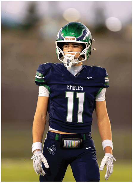 A person wearing a dark blue football uniform with the number eleven and a green helmet stands on a field with a focused expression. Description generated by AI