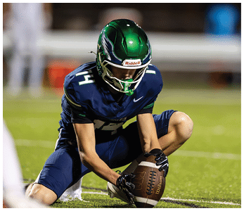 A person wearing a green helmet and navy blue sports uniform is kneeling on a football field while holding a football. Description generated by AI