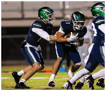 Two people in football uniforms with green helmets are engaged in a play on a football field, with one person handing off the ball to the other. Description generated by AI