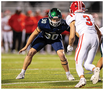 Two people in football uniforms are positioned on a field with one in a defensive stance wearing a dark blue uniform and helmet and the other in a white uniform with red stripes and the number three on the back. Description generated by AI