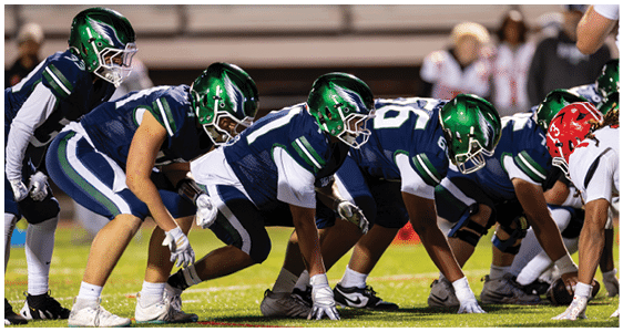 A group of football players in green helmets and navy uniforms are lined up on the field preparing for a play. Description generated by AI