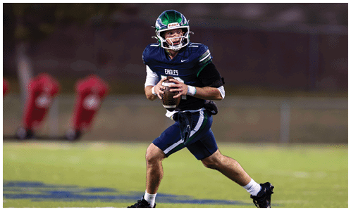 A person wearing a football uniform and helmet is running on a field while holding a football, appearing to prepare to throw it. Description generated by AI