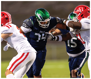 Two people in dark blue football uniforms with green helmets are blocking two people in white football uniforms with red helmets during a game. Description generated by AI