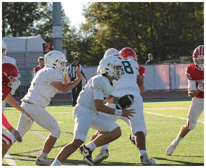 Several people in football uniforms are actively engaged in a play on a sunlit field with trees and a fence in the background. Description generated by AI