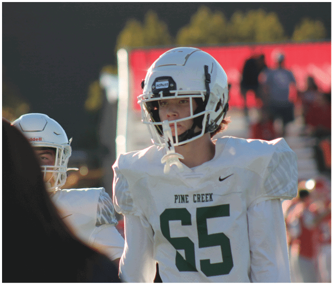 Two people wearing white football helmets and jerseys with the number 65 and the text Pine Creek are on a field with a blurred person in the foreground. Description generated by AI