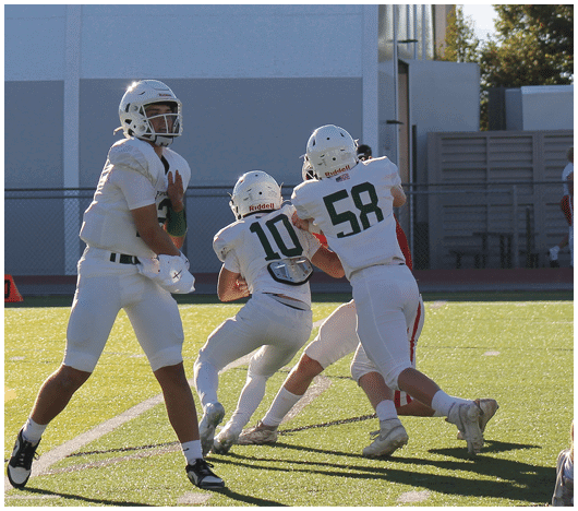 Several people in white football uniforms and helmets are actively engaged in a play on a sunlit football field near a building. Description generated by AI
