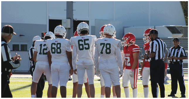 Players in white and red football uniforms stand on a field with referees nearby, preparing for a play. Description generated by AI