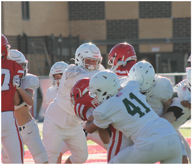 Several people wearing football helmets and uniforms are engaged in a play on a sunlit field with a building in the background. Description generated by AI