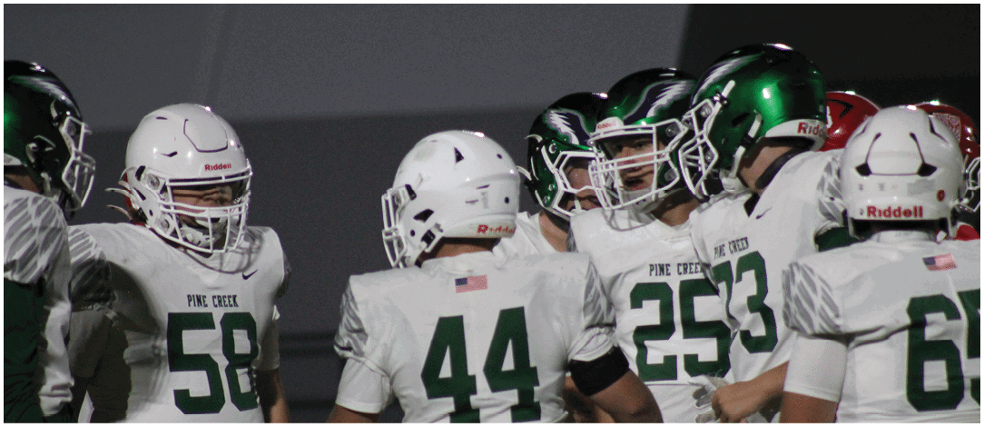 A person wearing a green helmet and white football uniform is holding a football while lying on the ground with two other players nearby on a football field. Description generated by AI
