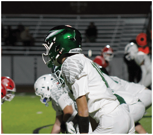People in football uniforms are lined up on a field preparing for a play during a game at night. Description generated by AI