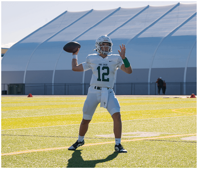 A person wearing a white football uniform with the number 12 is preparing to throw a football on a field with a large white dome structure in the background. Description generated by AI