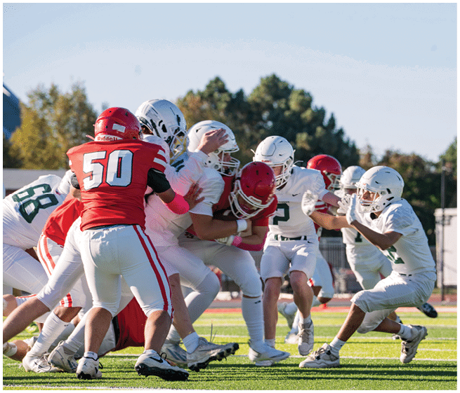 A group of people wearing football uniforms in red and white are engaged in a play on a sunny outdoor field with trees in the background. Description generated by AI