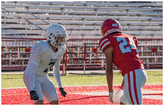 Two people in football uniforms are facing each other on a field with red and white markings, appearing ready for a play. Description generated by AI