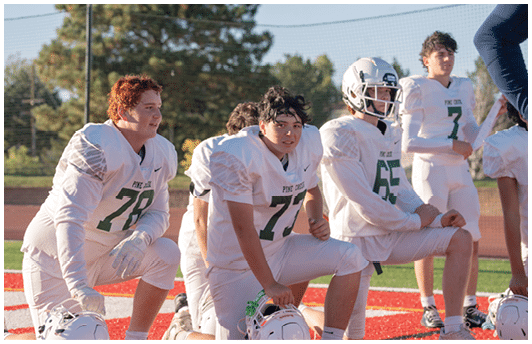 Four people in white football uniforms with green numbers are kneeling on a red field, with one person wearing a helmet and others holding helmets. Description generated by AI