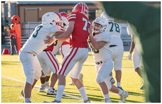 People wearing football uniforms are engaged in a play on a sunlit field with some players blocking and others preparing to move. Description generated by AI