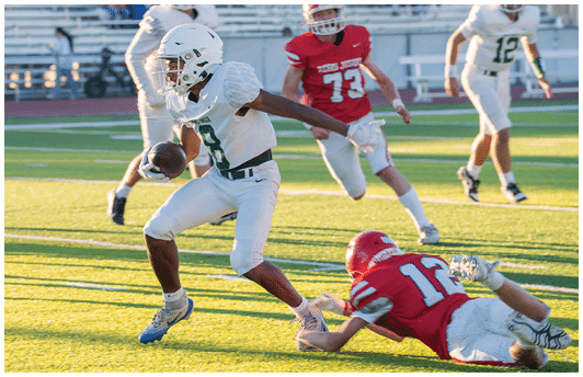 A person in white football gear is running with the ball while two people in red uniforms and two others in white uniforms are on the field during a game. Description generated by AI