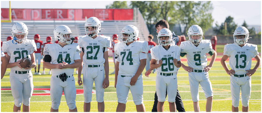 Five people wearing white football uniforms with helmets stand on a green field with a coach or adult in casual clothing behind them. Description generated by AI