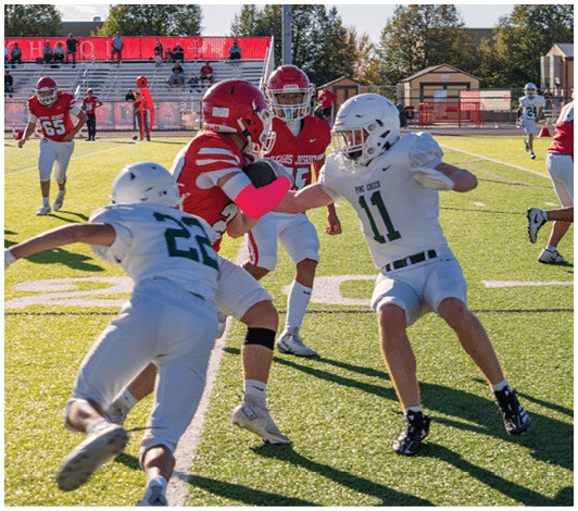 Two people in white football uniforms are attempting to tackle a person in a red football uniform who is holding the ball on a sunlit field with other players and spectators in the background. Description generated by AI