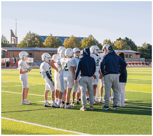 A group of people wearing football helmets and uniforms are gathered on a green field with two individuals in hoodies and sweatpants standing with them. Description generated by AI