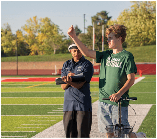 Two people stand on a sports field with one holding a clipboard and the other raising a hand while standing next to a scooter. Description generated by AI