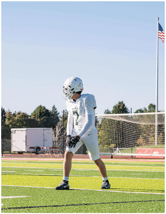 Two people in football uniforms are positioned on a field with a goalpost and trees in the background. Description generated by AI