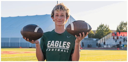 A person wearing a green Eagles football shirt is smiling and holding two footballs outdoors on a sports field with a large tent and bleachers in the background. Description generated by AI
