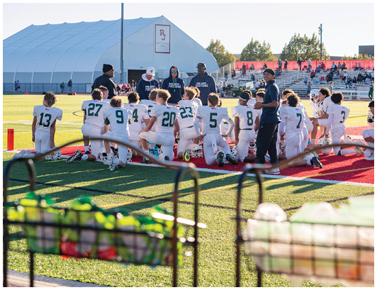 A group of people in football uniforms kneel on a field while two adults in sportswear stand nearby with bleachers and spectators in the background. Description generated by AI