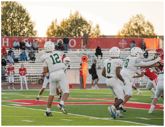 Players in white football uniforms are engaged in a play on a green field with a football mid-air, while players in red uniforms defend and spectators watch from the bleachers. Description generated by AI