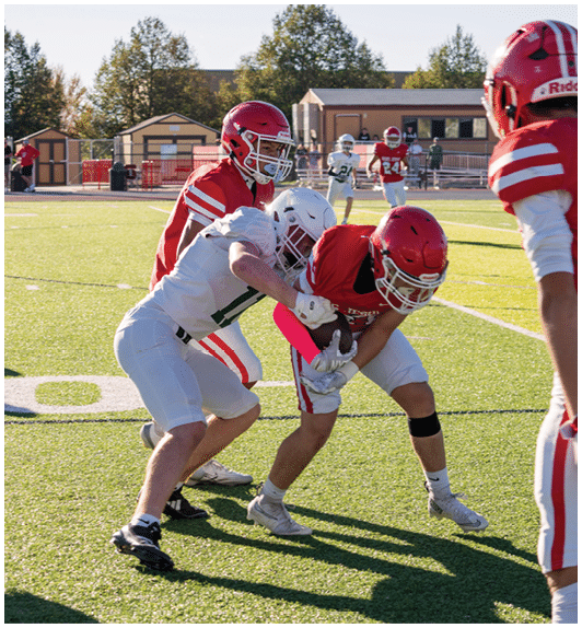 People wearing football uniforms are actively engaged in a game on a sunny field with trees and a building in the background. Description generated by AI