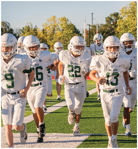 A group of young people wearing white football uniforms and helmets are running on a green field with trees in the background. Description generated by AI