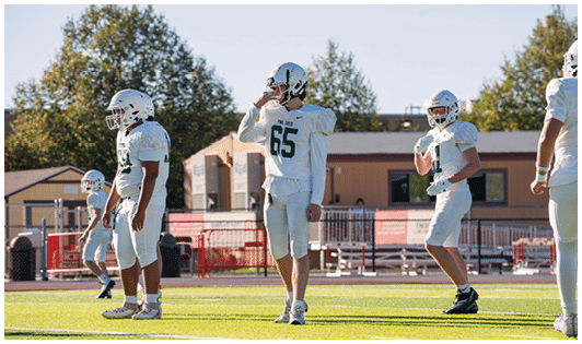 Several people in white football uniforms and helmets stand on a green field with trees and buildings in the background. Description generated by AI