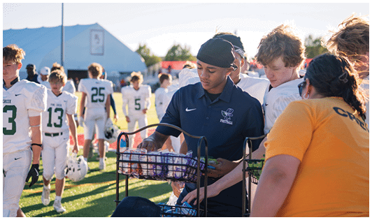 People in sports uniforms gather on a field while one person in a dark polo shirt arranges items in a basket. Description generated by AI
