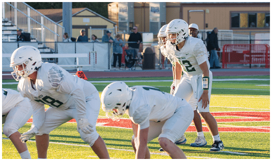 A group of people wearing white football uniforms and helmets are positioned on a football field preparing for a play during a game. Description generated by AI