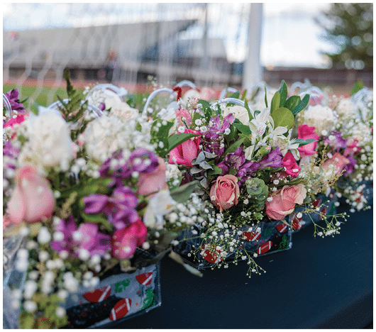 Colorful floral arrangements with pink, white, and purple flowers are placed in decorated containers on a dark tablecloth outdoors near a sports field. Description generated by AI