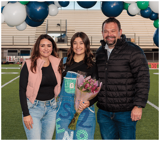 Three people smiling and standing close together under blue and white balloons with one person holding a bouquet of flowers. Description generated by AI