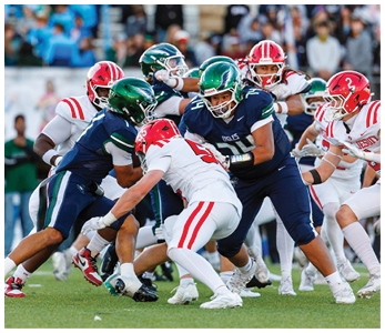 Several people wearing football uniforms in blue and white with helmets are engaged in a close, intense play on a grassy field during a game. Description generated by AI