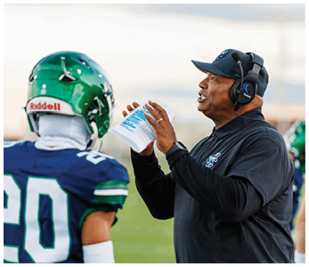A person wearing a headset and black sportswear is holding a play sheet and speaking to a person in a green football helmet and blue jersey with the number 20 on a field. Description generated by AI