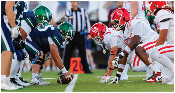 Two opposing American football teams in helmets and uniforms are lined up facing each other on a field with a referee standing in the background. Description generated by AI